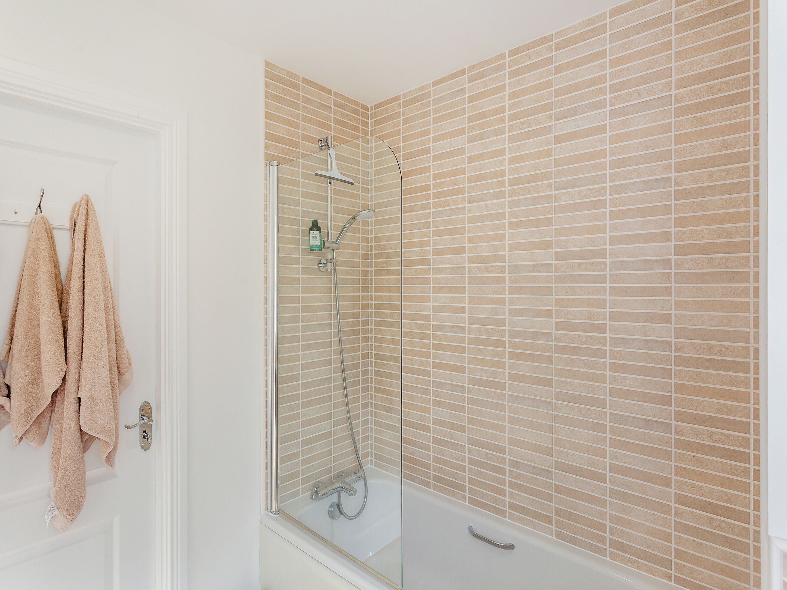 Ein Badezimmer mit Duschkabine, beige gefliesten Wänden und einer weißen Tür, an der zwei Handtücher hängen.
