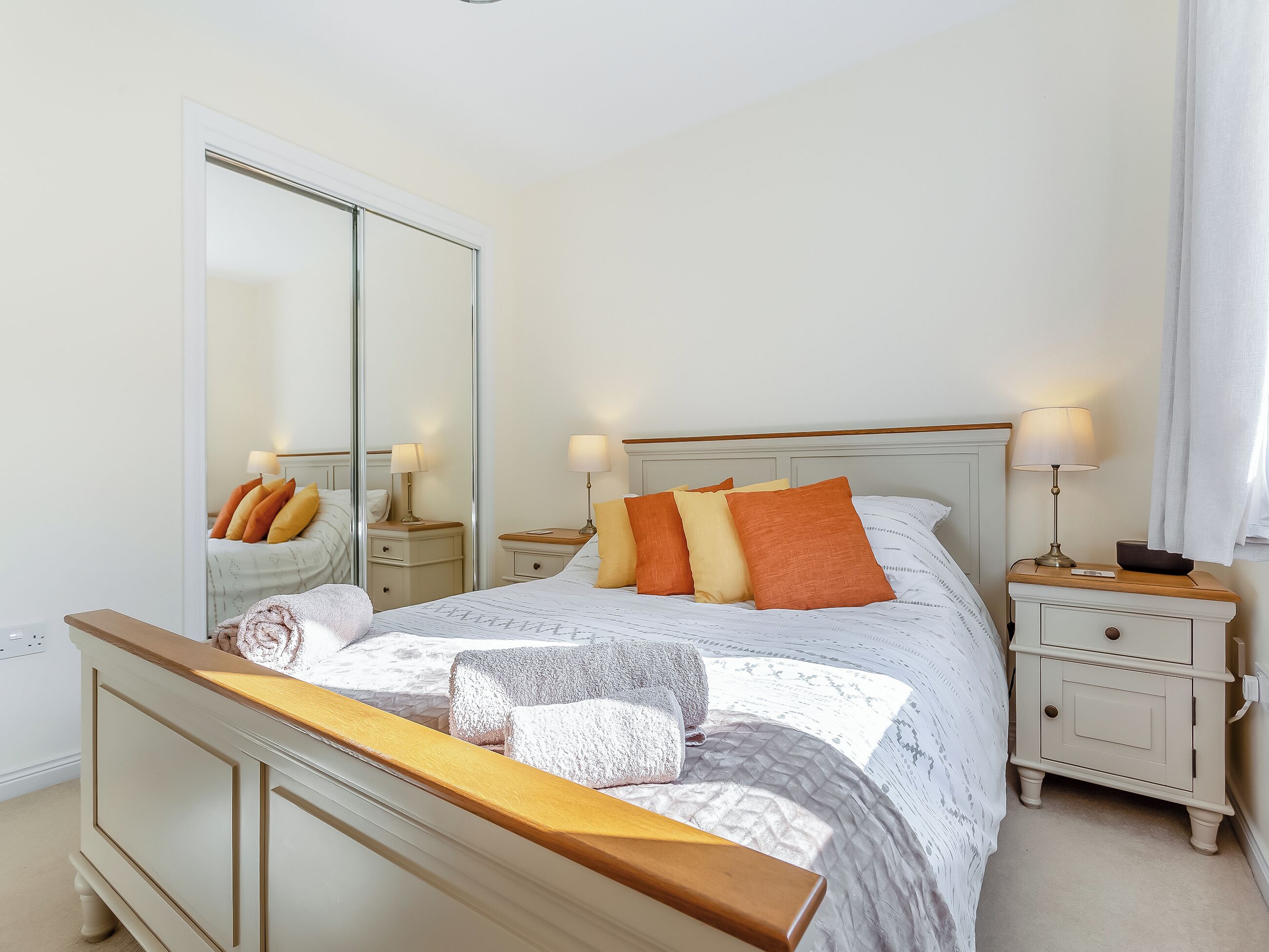 A bedroom with a large mirror, a bed with multiple pillows, bedside tables with lamps, and a wooden headboard.