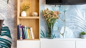 Un salon doté d’un téléviseur à écran plat fixé au mur, d’étagères en bois ornées de livres et d’objets décoratifs, et d’un vase rempli de fleurs posé sur une commode blanche.