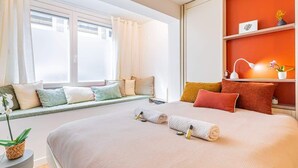 Une chambre d’hôtel avec un grand lit, un coin fenêtre agrémenté de coussins, une table de chevet avec une lampe et une étagère avec des objets décoratifs.