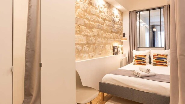 Un dormitorio moderno con pared de piedra, una cama con almohadas coloridas, una mesita de noche y una ventana con cortinas.