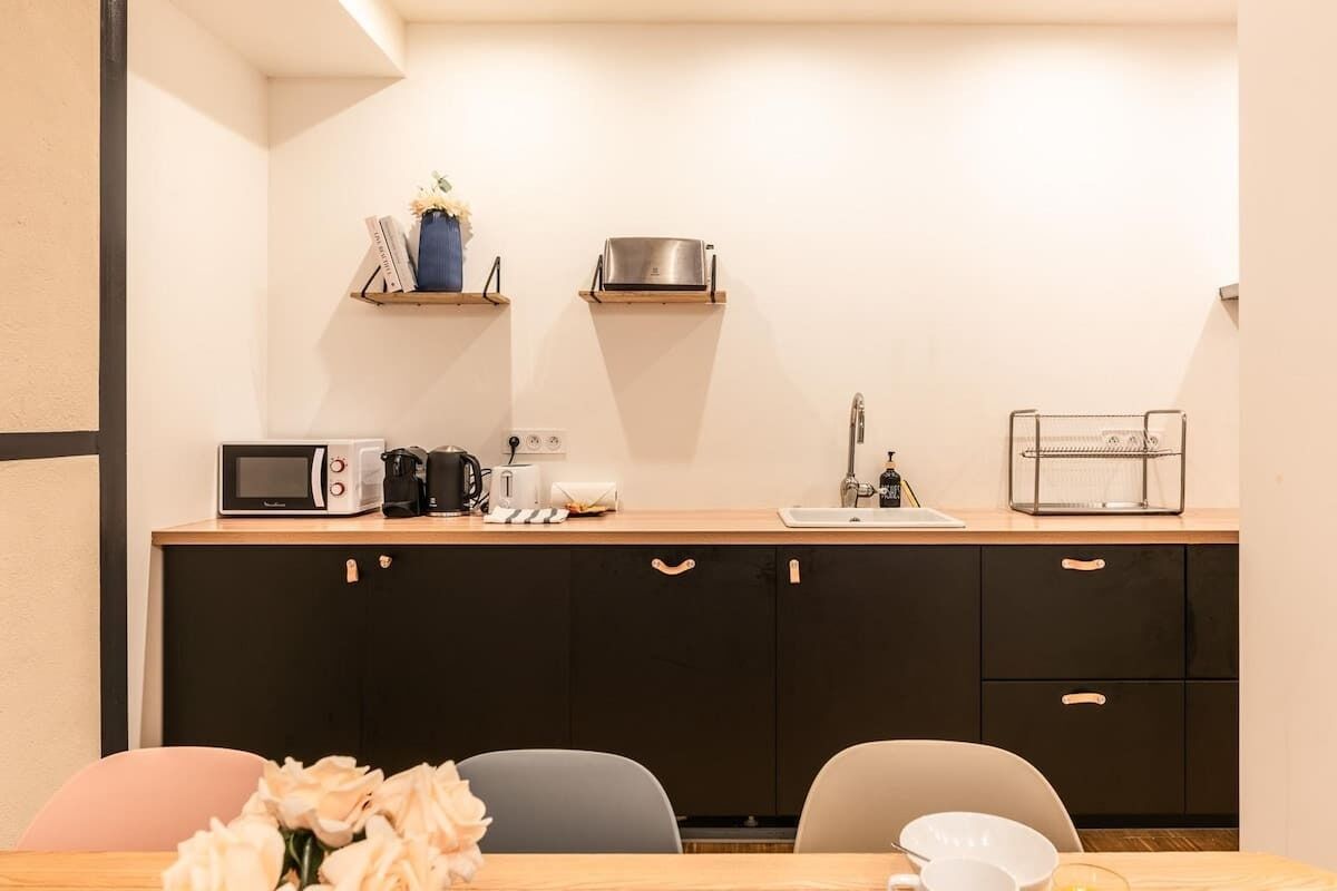 A compact kitchen with a microwave, coffee maker, toaster, and a sink. There are two chairs and a table with a vase of flowers.