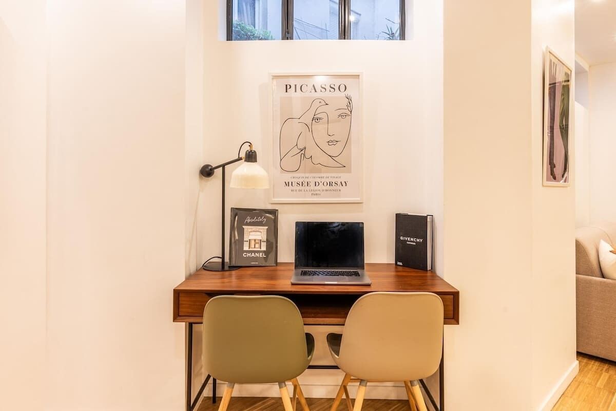 Een moderne werkplek met een houten bureau, twee stoelen, een laptop, een lamp en een Picasso kunstwerk in een lijst.