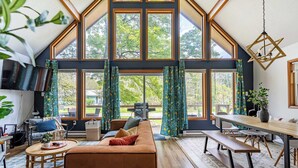A modern interior with a high ceiling, large windows, a sofa, a dining table, and a bench. - Blue Diamond Hot Tub Fire Pit 3 BDR (Tobyhanna)