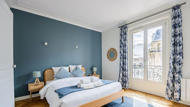 Un dormitorio con cama, cabecera de madera, dos mesitas de noche con lámparas, alfombra azul y un ventanal con cortinas.