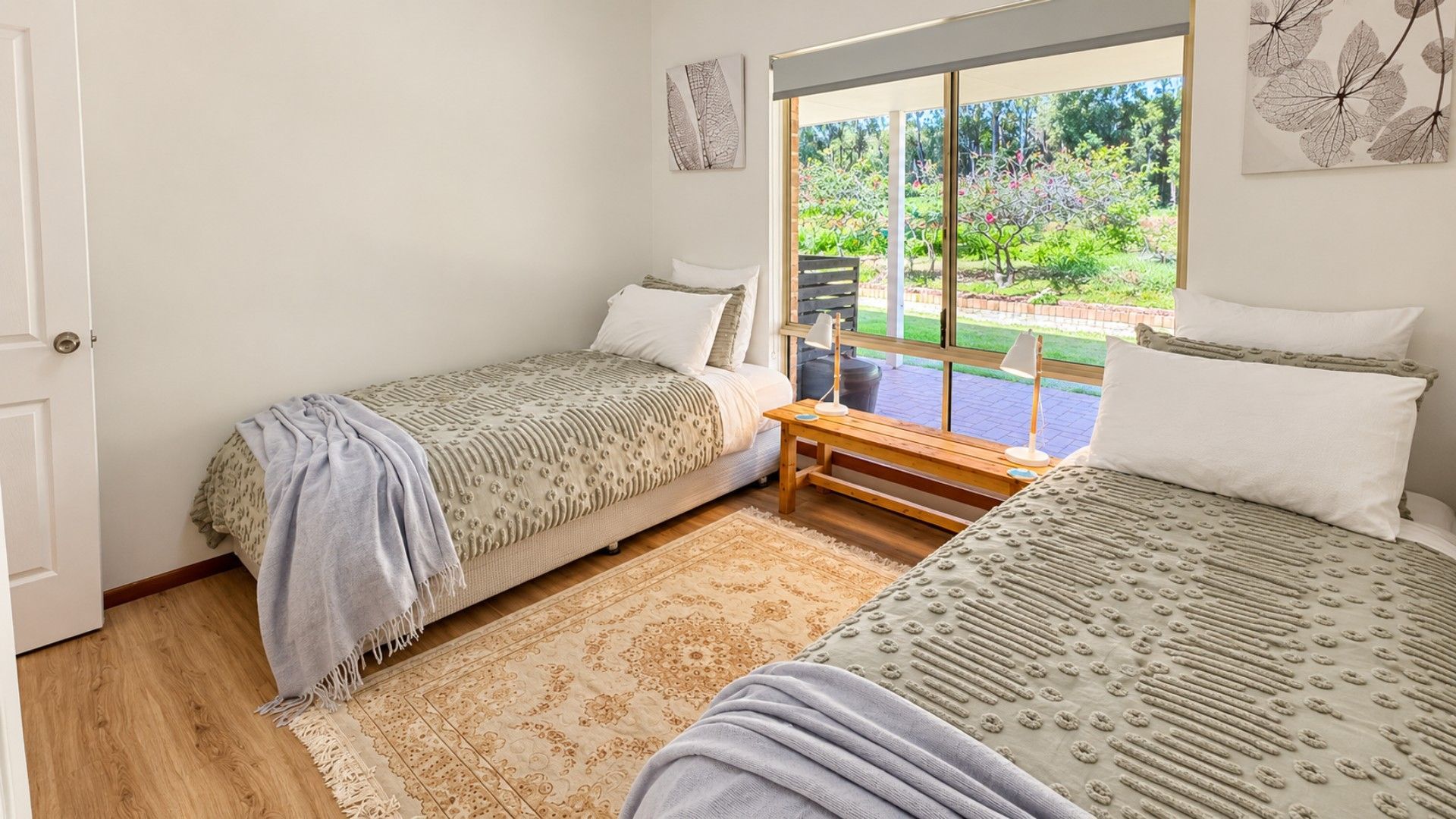 A bedroom with two single beds, a bench, a window with a view of a garden, and a door.
