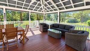 Una terraza techada al aire libre con piso de madera, muebles de mimbre y una mesa de vidrio.