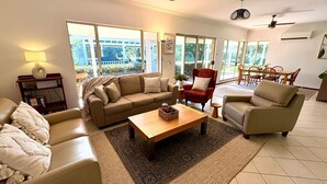 Amplia sala de estar con un sofá beige, una mesa de centro de madera y un sillón rojo. Amplias ventanas ofrecen vistas al exterior.