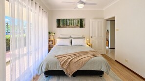 Un dormitorio con cama, ventilador de techo, ventana con cortinas, un cuadro en la pared y una puerta que da a otra habitación.