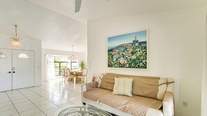 Ein geräumiges Wohnzimmer mit einem weißen Sofa, einem Glastisch und einem Bild einer Küstenstadt an der Wand.
