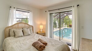 Ein Schlafzimmer mit einem großen Bett, einem Nachttisch mit Lampe und Blick auf einen Swimmingpool durch eine Glastür.
