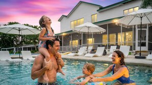 Una piscina con sillas de descanso y sombrillas, una casa con grandes ventanales y un cielo al atardecer.
