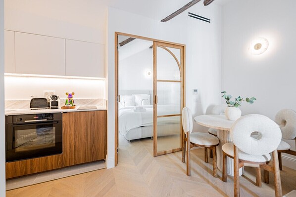 Una cocina moderna con lavavajillas, horno y armarios de madera. Hay una zona de comedor con una mesa redonda y sillas, y una cama con ropa de cama blanca al fondo.