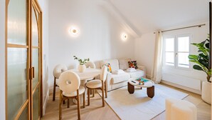 A spacious living room with a high ceiling, a dining area with a round table and chairs, a sofa set, and a large window providing natural light.
