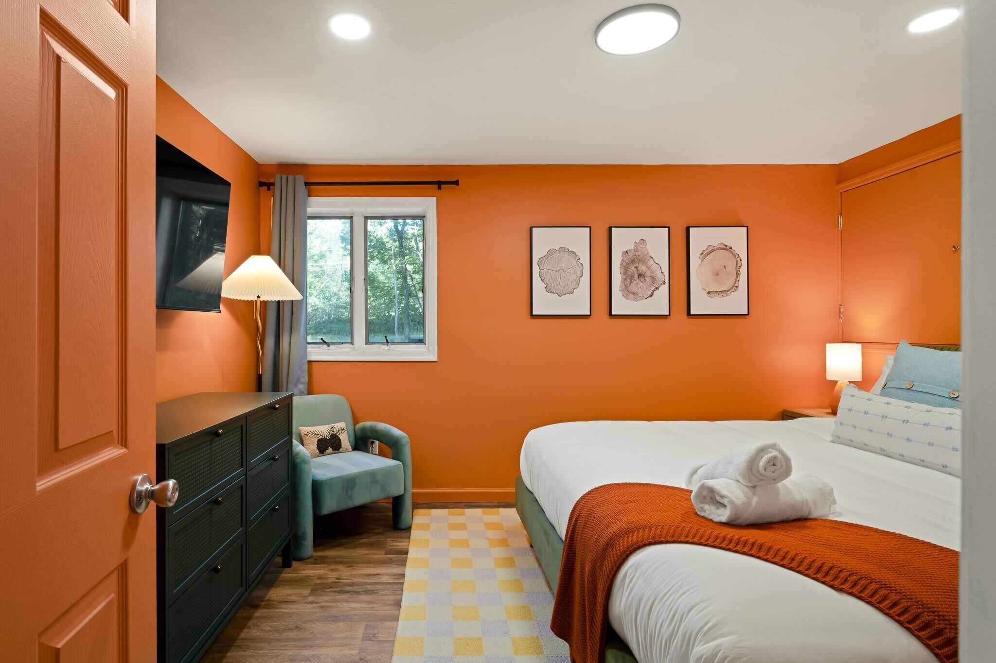 A bedroom with an orange wall, a bed with white bedding, a blue chair, a dresser, and three framed pictures on the wall.