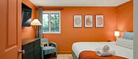 A bedroom with an orange wall, a bed with white bedding, a blue chair, a dresser, and three framed pictures on the wall.