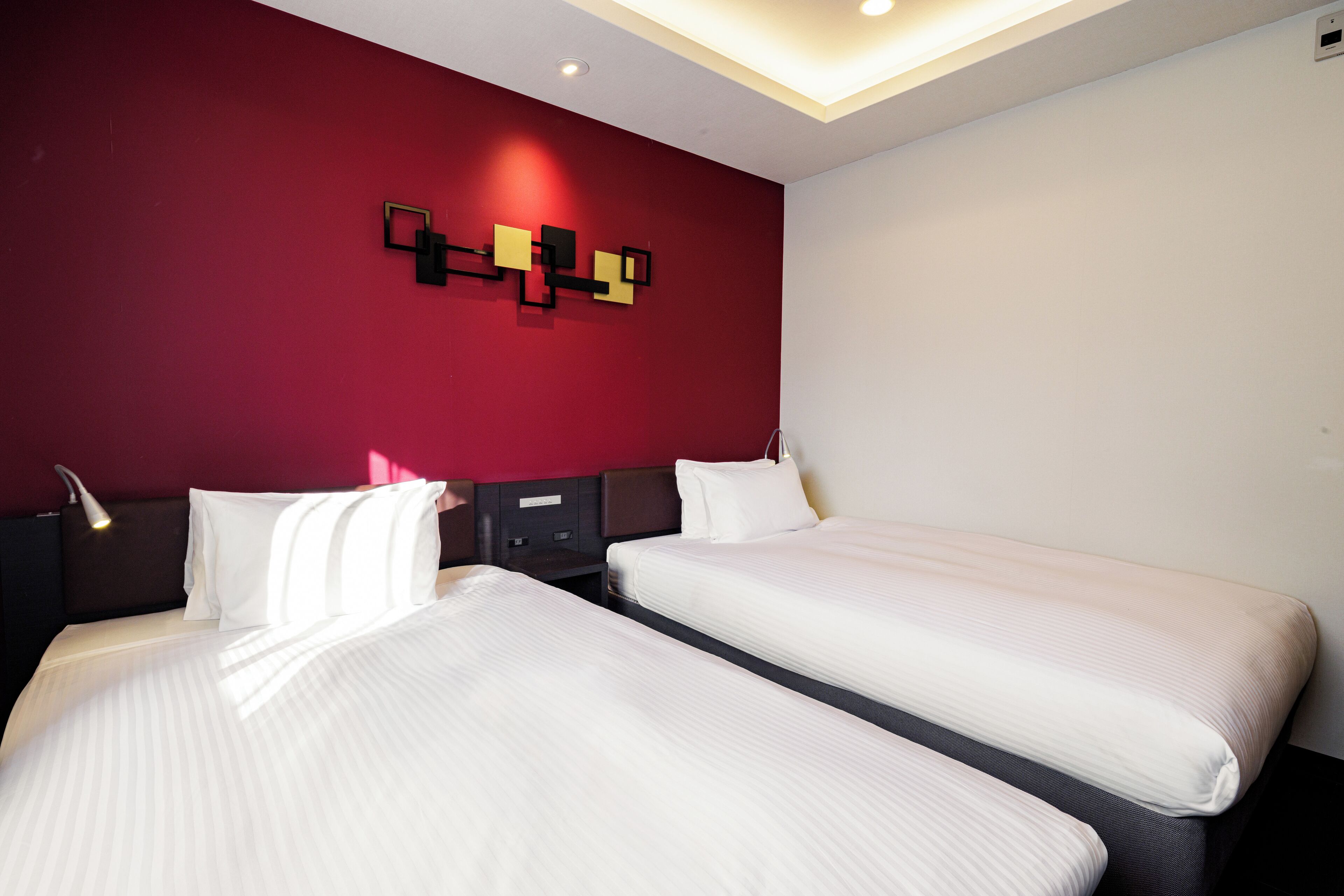 A hotel room with two beds, a red accent wall, and a modern light fixture.