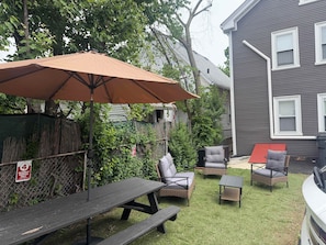 A backyard with a picnic table, two wicker chairs, a small table, and an umbrella.