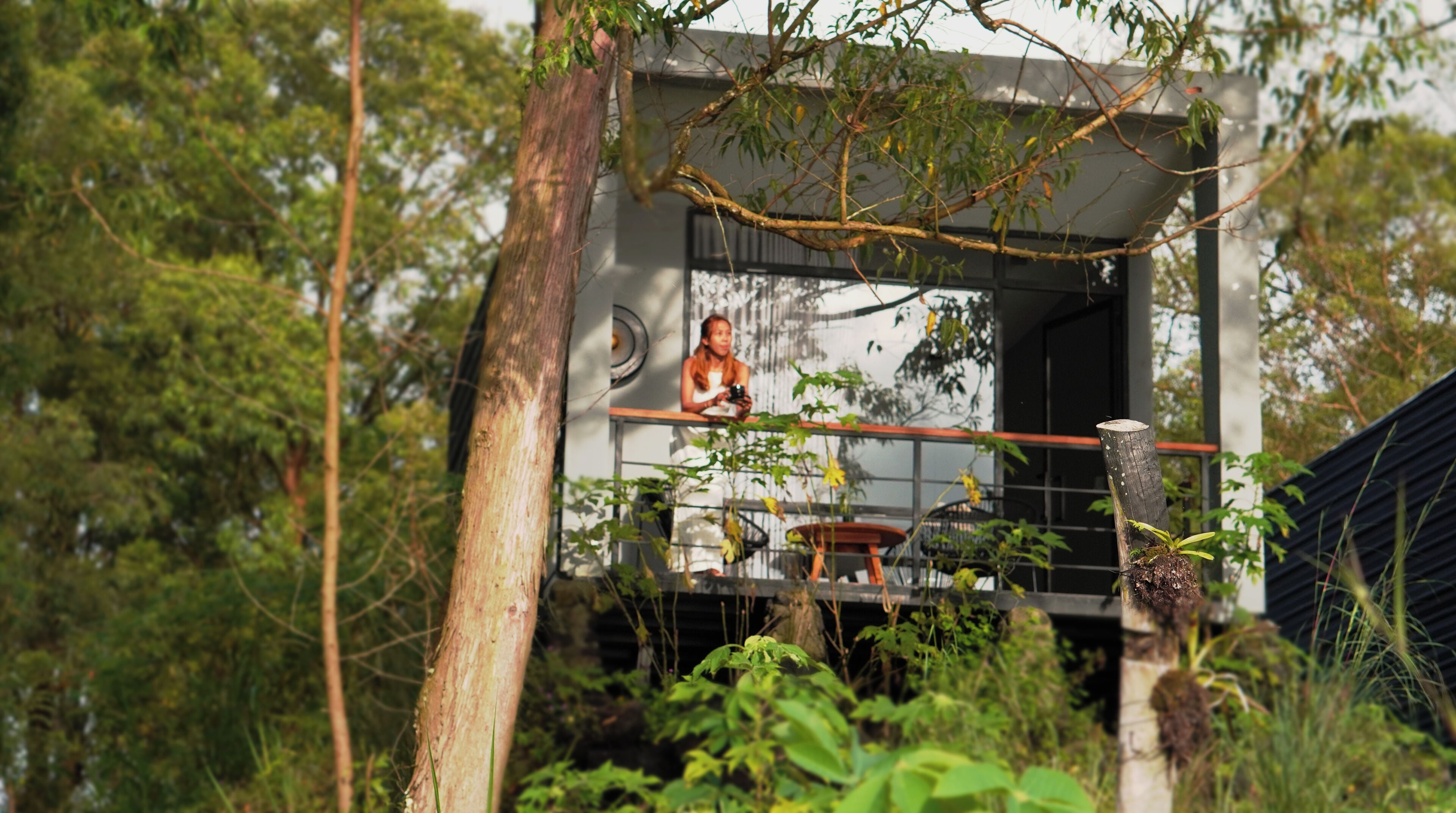 A balcony with a railing, a person seated, and a view of trees and a building.