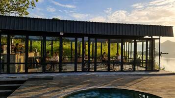 Restaurante con terraza al aire libre y vistas a un cuerpo de agua, que cuenta con una cubierta de madera y cerramiento de vidrio.