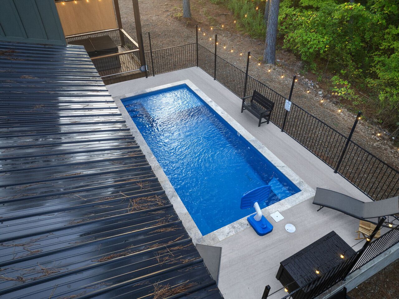 Une piscine rectangulaire à l’intérieur bleu, entourée d’une terrasse en béton et d’une clôture en métal noir. On y trouve un banc et une petite table près de la piscine.