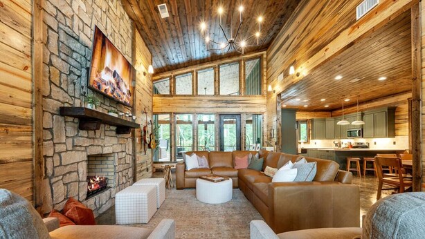 A cozy log cabin interior with a stone fireplace, leather sofas, and a large window overlooking greenery. - Sleeps 18 people with a private heated pool, hot tub, playground, fire pit, and a private arcade! (Broken Bow)