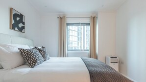 Un lit bien fait, avec une couette blanche et des oreillers à motifs, une grande fenêtre avec des rideaux et un climatiseur fixé au mur.