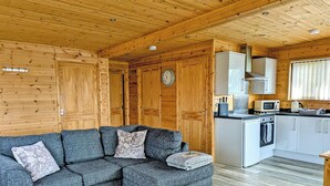 Un intérieur de chalet chaleureux avec un canapé d’angle, des murs en bois, une cuisine dotée de meubles blancs et un plafond en bois.