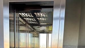 Un ascenseur à l’intérieur métallique, un panneau de commande avec un bouton rouge d’urgence et une vue sur l’extérieur à travers les portes en verre.