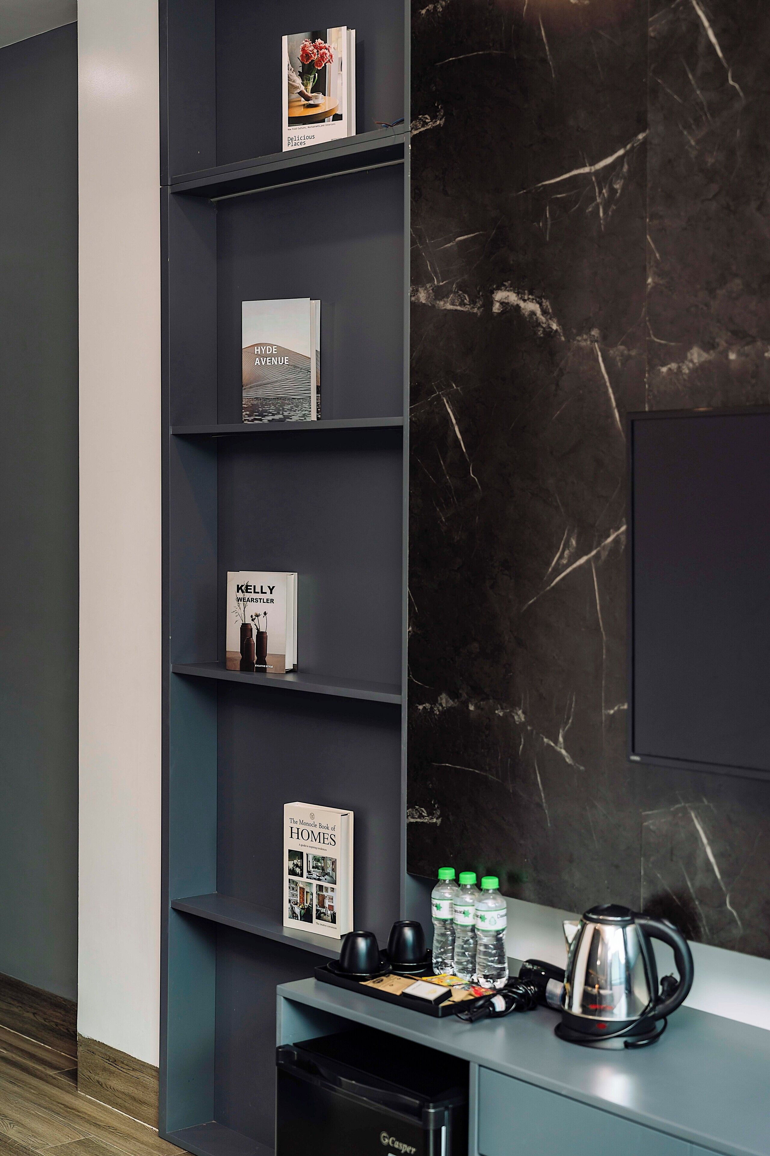 A modern interior with a marble wall, a black shelf unit displaying books, and a coffee station with a kettle and water bottles.