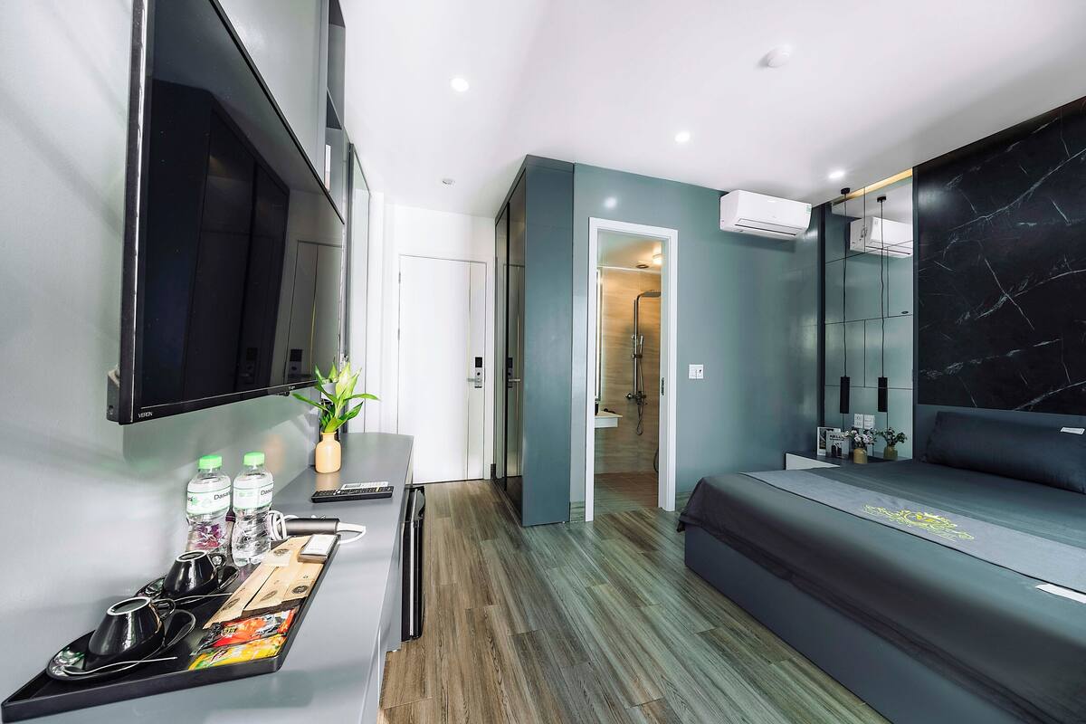 Une chambre d’hôtel moderne équipée d’un lit, d’un bureau avec un plateau contenant du thé et des bouteilles d’eau, d’une télévision et d’une salle de bain visible à travers une porte ouverte.
