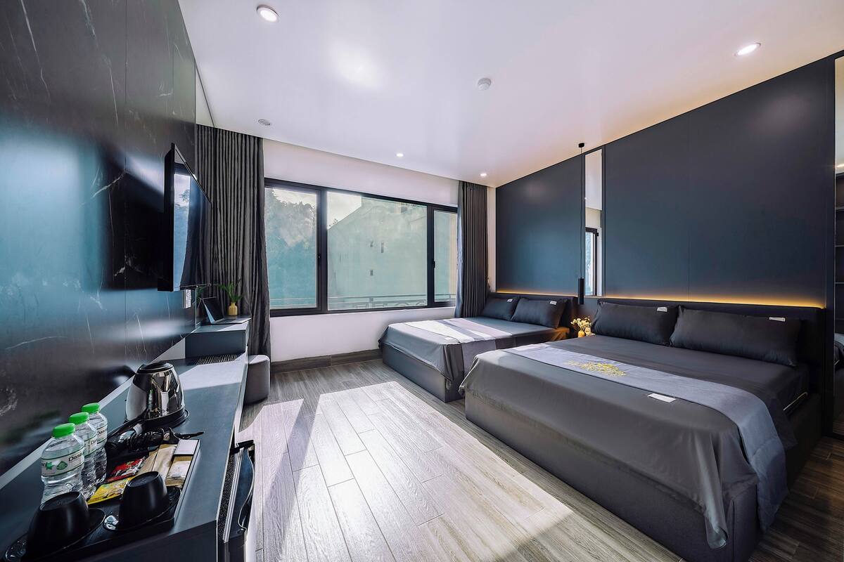 Une chambre d’hôtel moderne équipée d’un grand lit, d’un lit plus petit, d’un bureau avec une bouilloire et des bouteilles d’eau, et d’une grande fenêtre donnant sur l’extérieur.