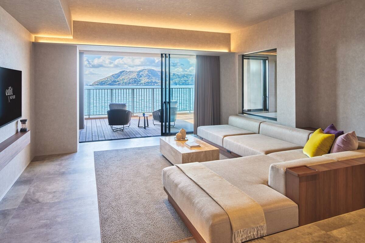 A modern living room with a sofa set, a coffee table, and a TV on the wall. There is a balcony with a view of the sea and mountains.