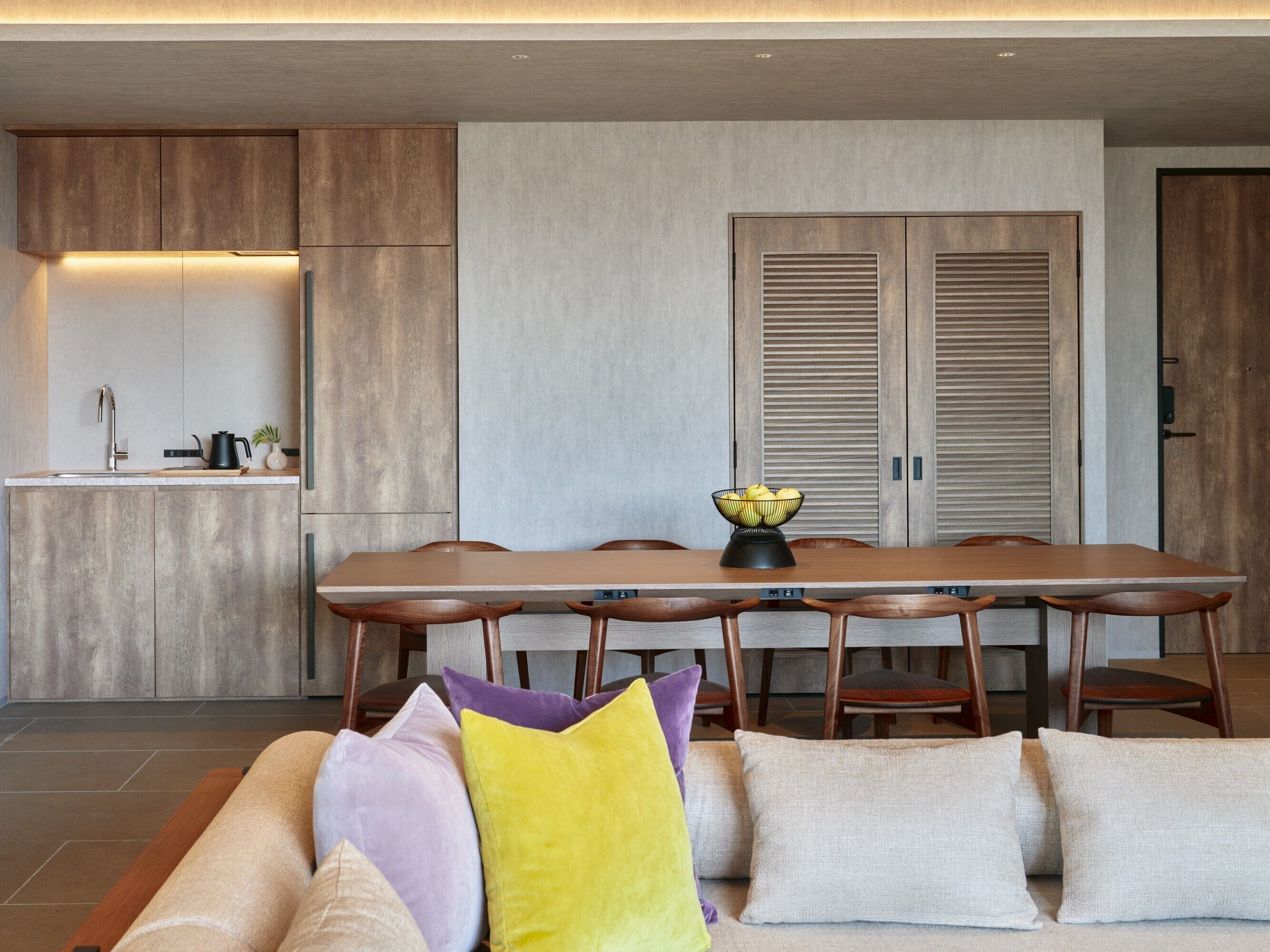 A modern kitchen with wooden cabinets, a dining table with chairs, and a sofa with cushions.