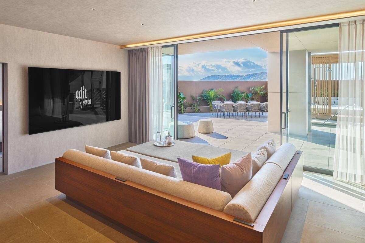 A modern living room with a large flat-screen TV, a sofa with cushions, a coffee table, and a balcony with outdoor seating.