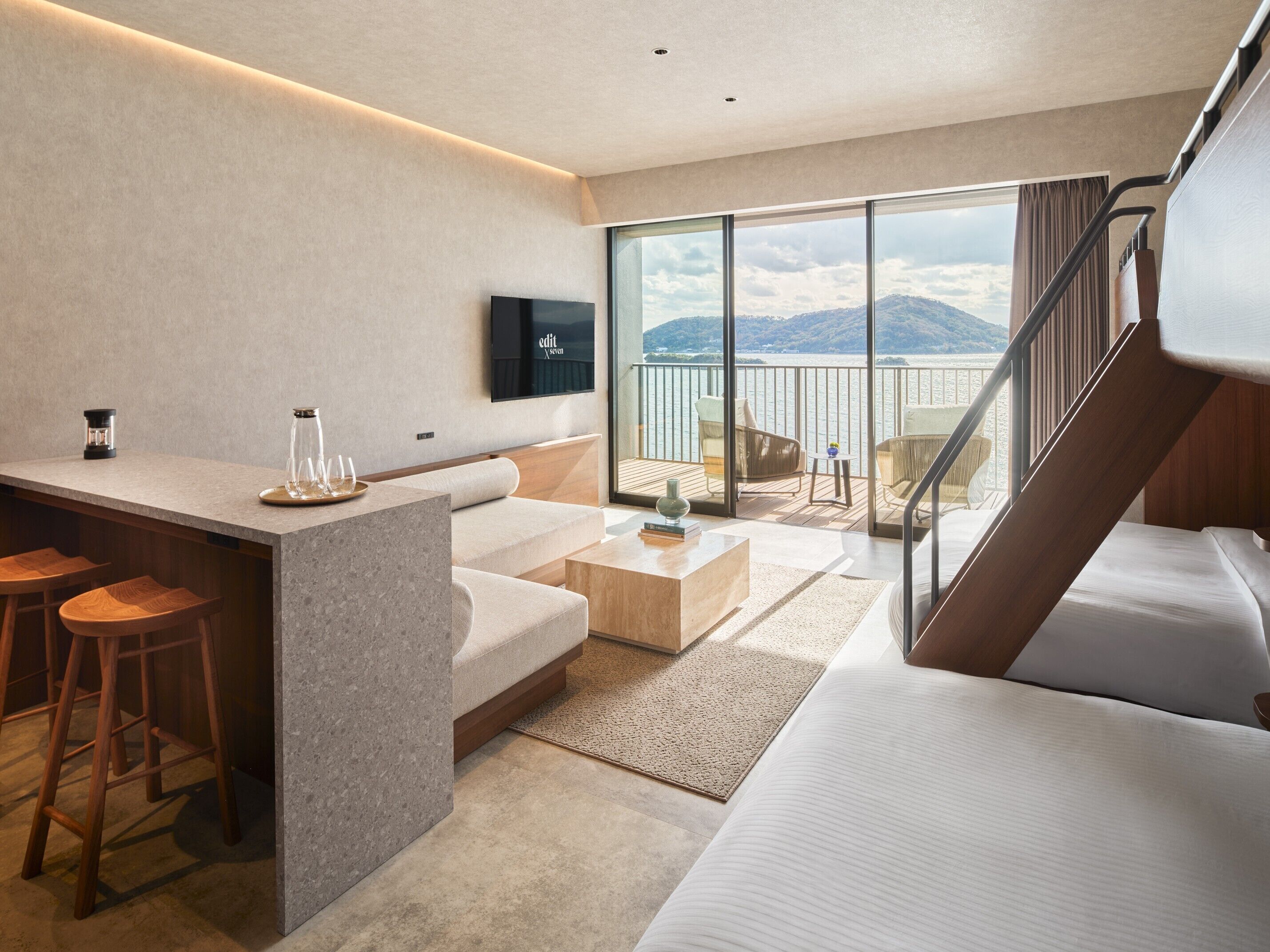 A modern hotel room with a balcony, a flat-screen TV, a kitchen area, and a large bed.