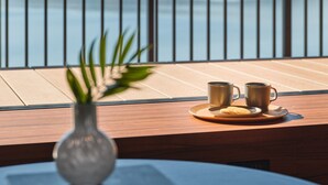 Une table en bois avec deux tasses et une assiette, un vase avec une plante, et une vue sur un lac et des montagnes à travers des barreaux métalliques.