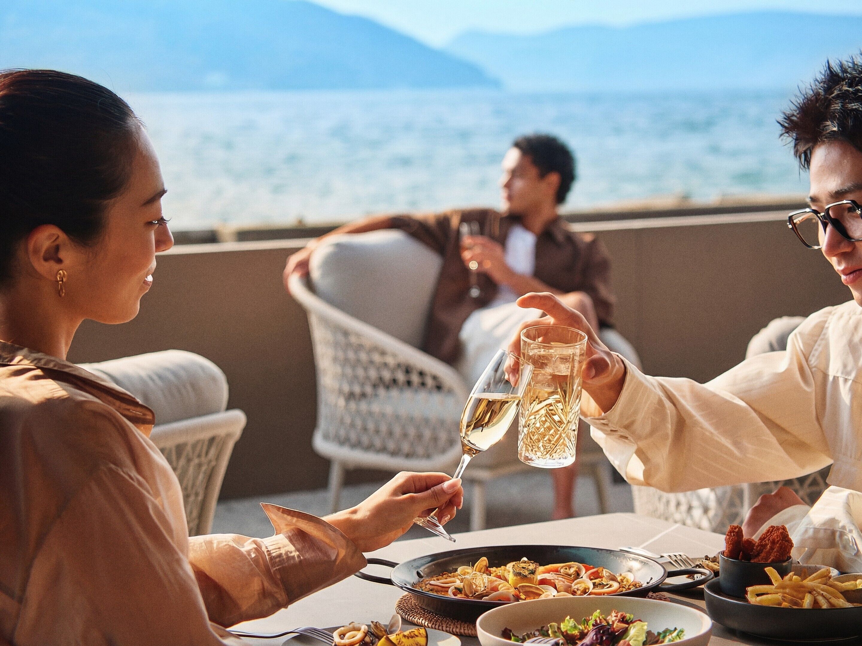 Un coin repas avec une table dressée pour le dîner, une vue sur la mer et une personne qui porte un toast avec un verre.