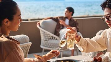 Un coin repas avec une table dressée pour le dîner, une vue sur la mer et une personne qui porte un toast avec un verre.
