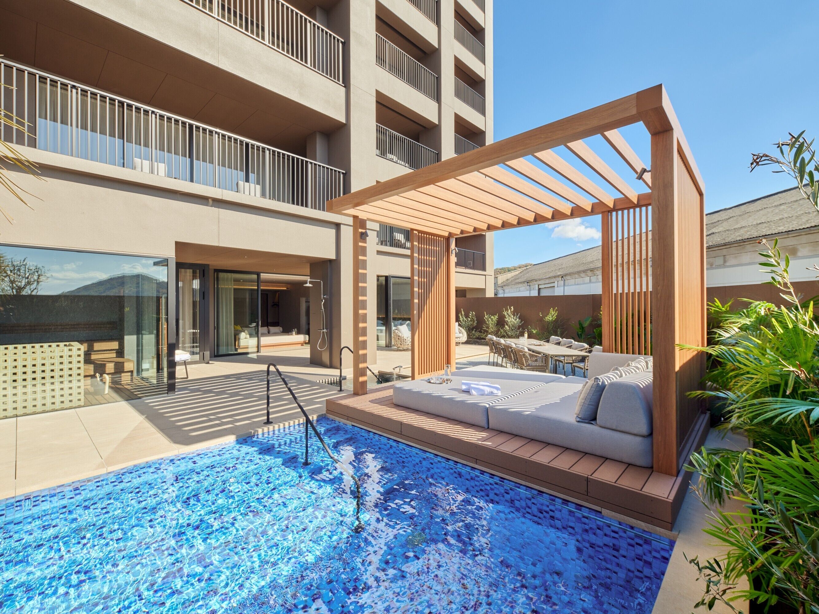 A modern outdoor pool area with a wooden pergola, seating, and a view of a high-rise building.