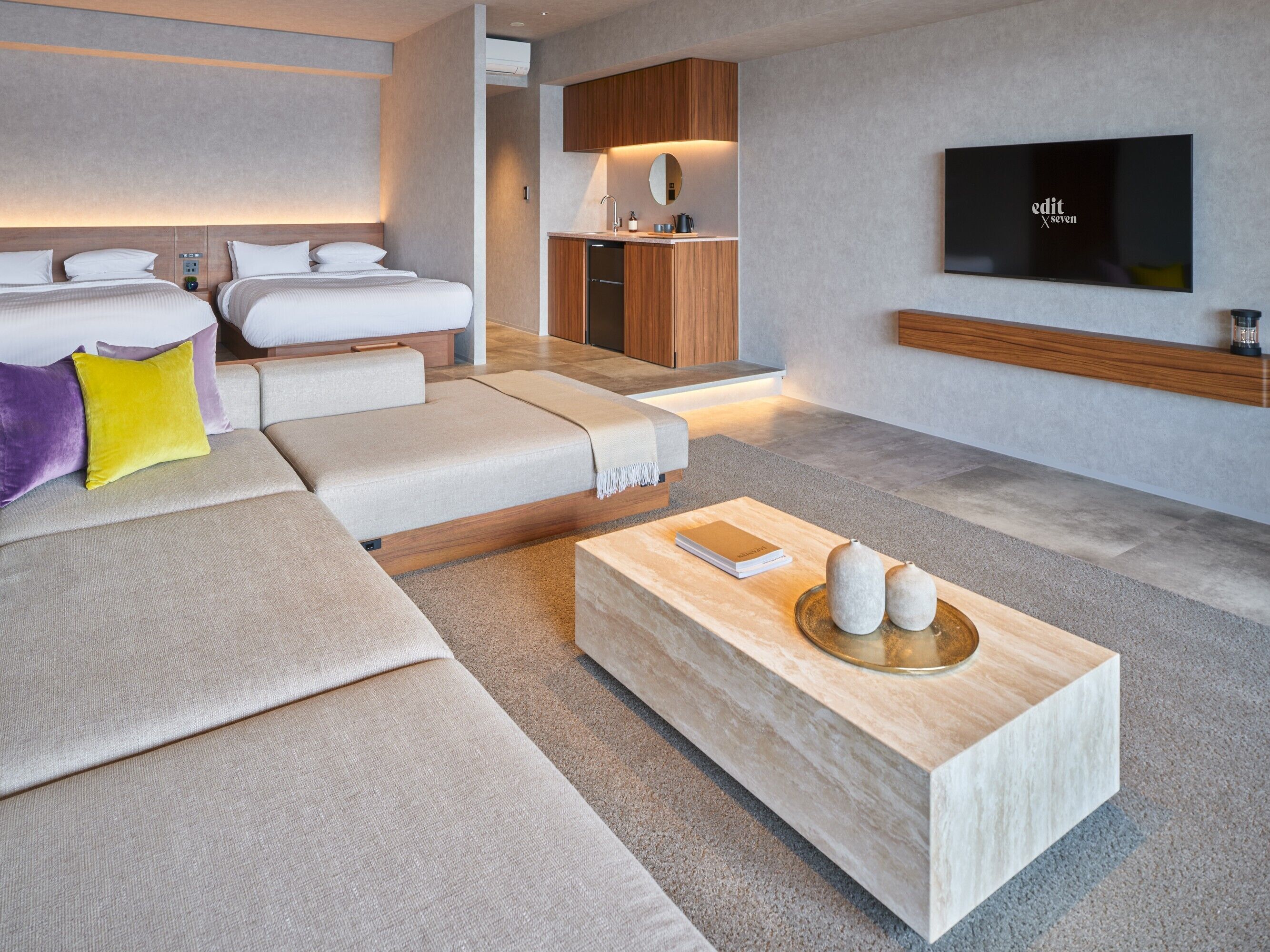 A modern hotel room with a large sofa, a small kitchen area, and a flat-screen TV mounted on the wall.