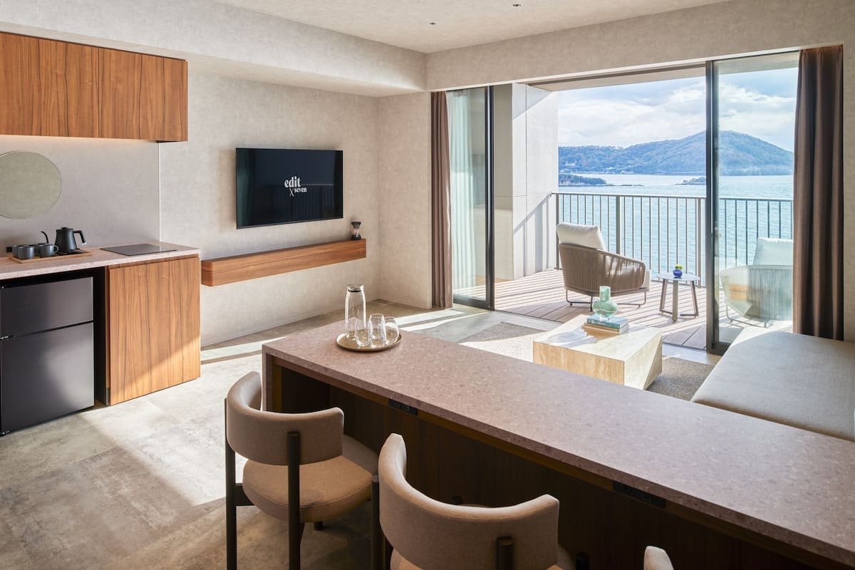 A modern hotel room with a balcony, a flat-screen TV, a dining area with chairs, and a view of the sea.