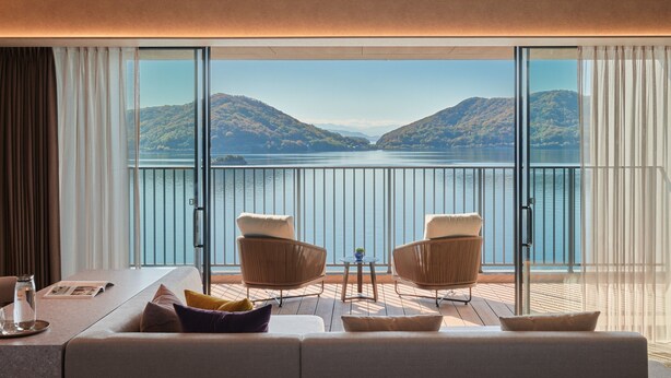 Une chambre d’hôtel avec un balcon donnant sur un lac et des collines. On y trouve un canapé, deux fauteuils et une petite table avec un verre et un magazine.