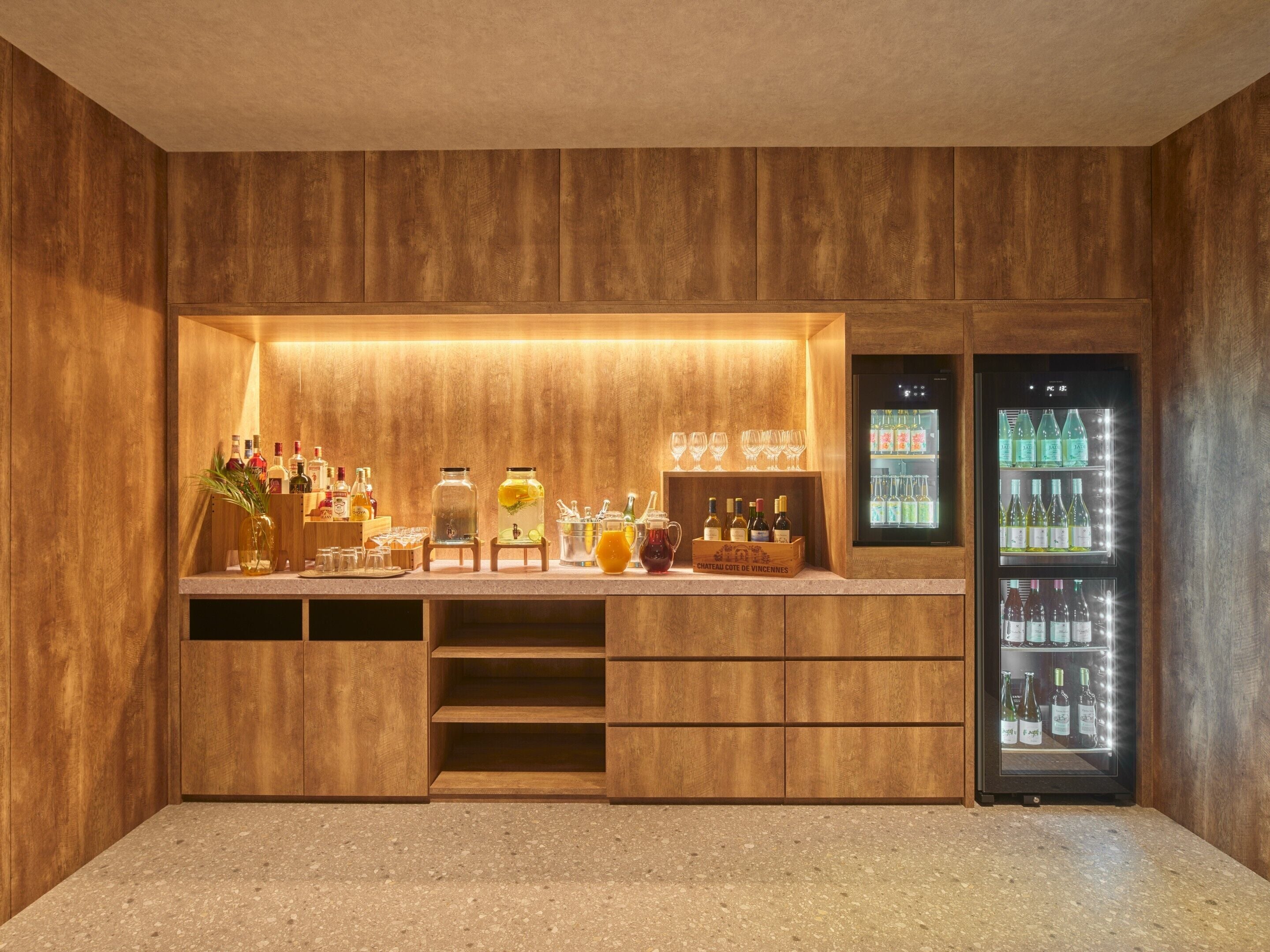 Een bar met houten lambrisering, een aanrecht met diverse flessen en glazen, en een koelkast vol drankjes.