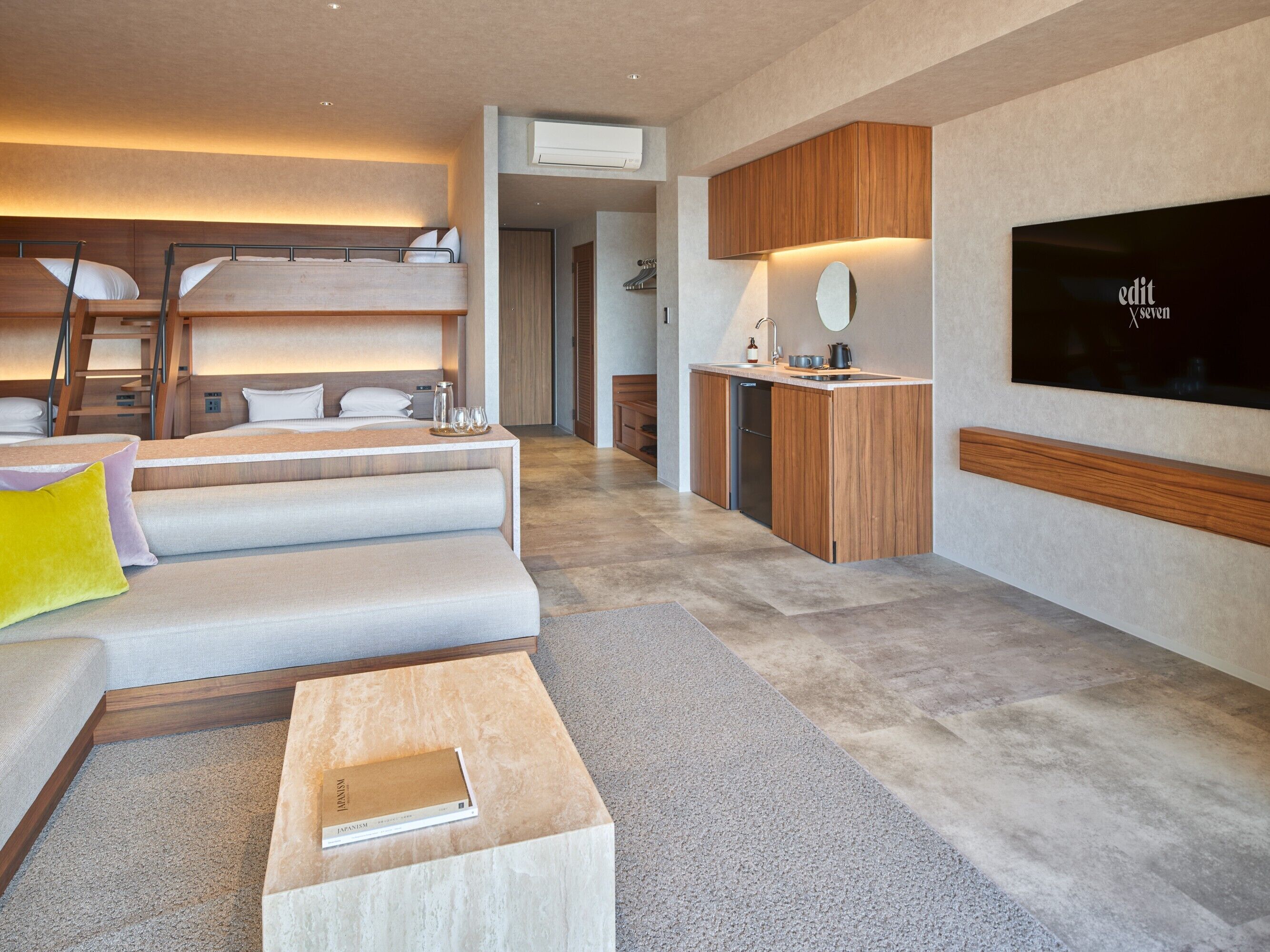 A modern hotel room with a bunk bed, sofa, flat-screen TV, and a small kitchenette.