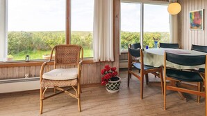 A room with a dining table, chairs, a wicker chair, a potted plant, and large windows with a view of greenery. - 6 Person Holiday Home in Lundby (Lundby)