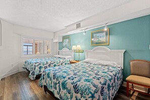 A bedroom with two beds, a window with shutters, a chair, and a painting on the wall.