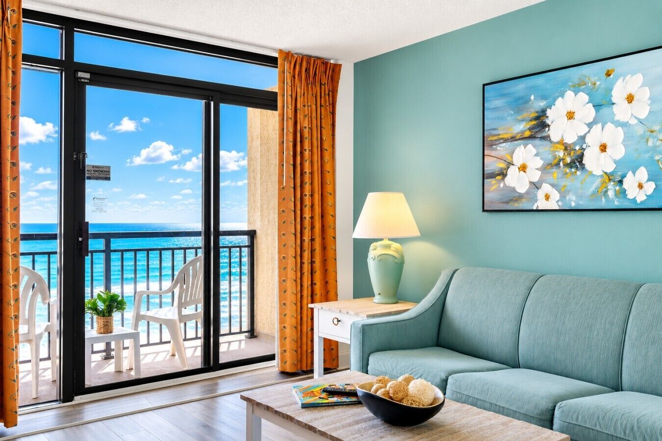 A room with a view of the ocean, a sofa, a table with a bowl of snacks, a lamp, and a painting of flowers.