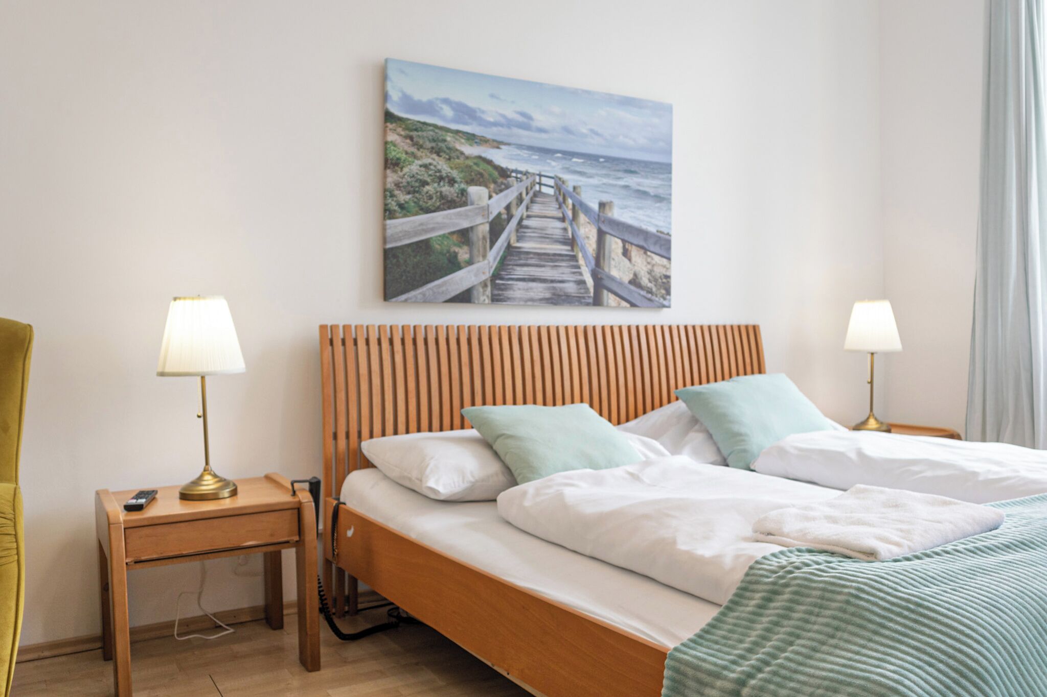 Un dormitorio con cabecera de madera, dos mesitas de noche con lámparas, cama con ropa de cama y almohadas blancas, y un cuadro de una escena costera en la pared.