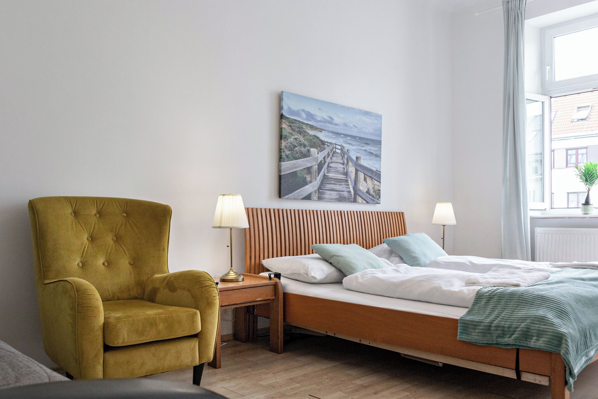 Une chambre à coucher avec un lit en bois, un fauteuil vert, une table de chevet avec des lampes et un tableau représentant un paysage côtier.